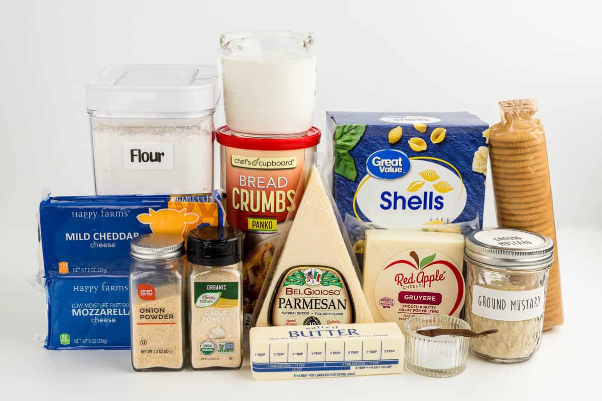 Flour, cheese, onion powder, garlic powder, butter, bread crumbs, salt, ground mustard, crackers, and pasta on a counter surface.