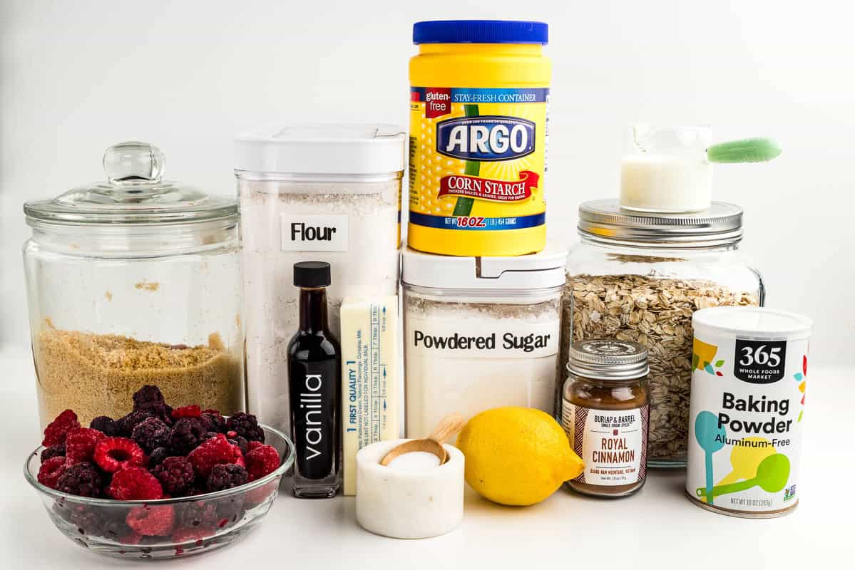 Berries, brown sugar, vanilla, flour, salt, lemon, cinnamon, corn starch, powdered sugar, butter, oats, baking powder, and milk on a countertop.