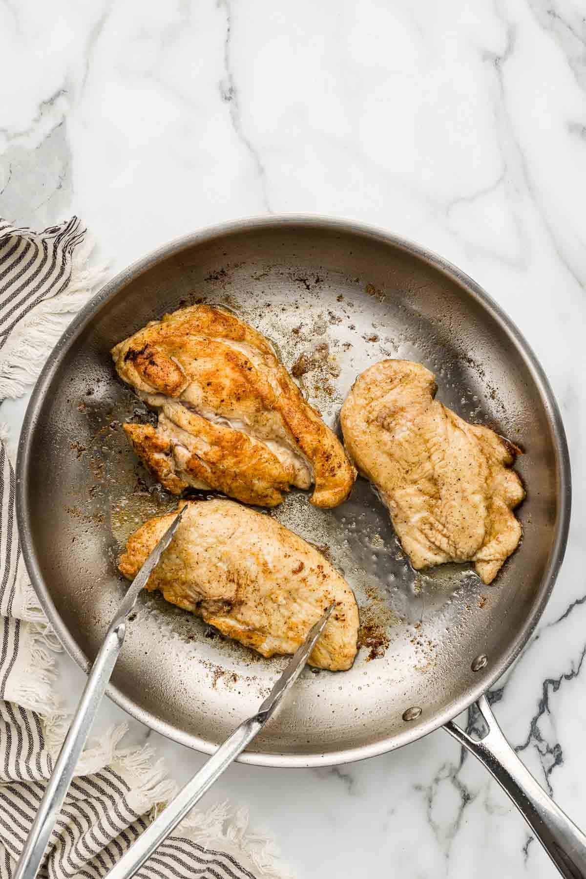 A pair of tongs over a skillet of cooking chicken.