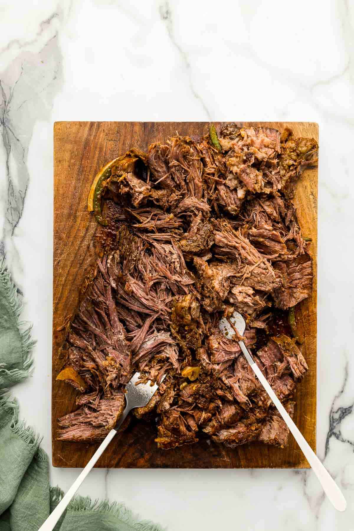 Beef being shredded with two forks on a wooden cutting board.