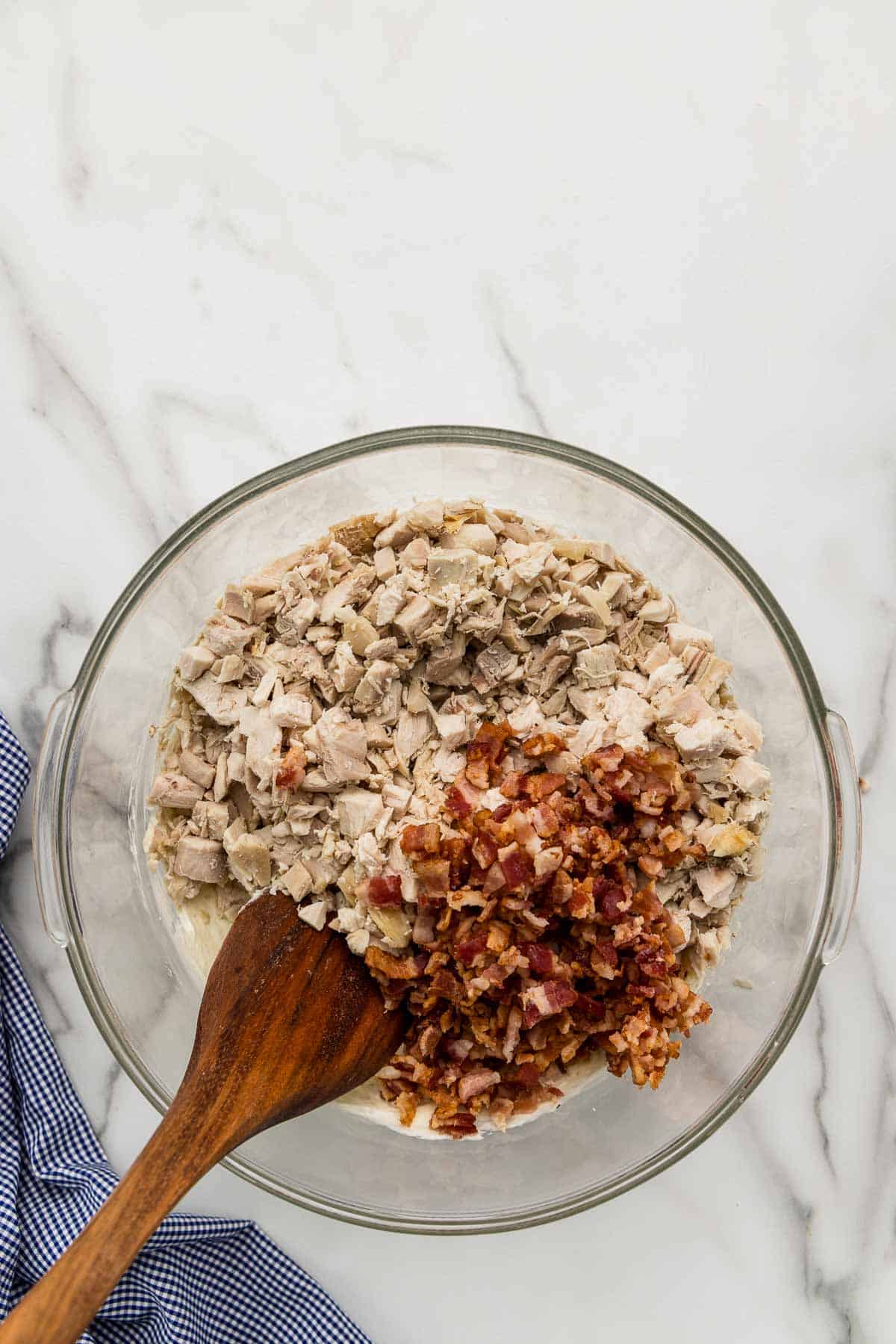 Chicken and bacon in a glass mixing bowl with a wooden spoon.