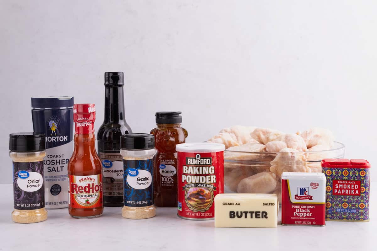 Hot sauce, spices, salt, baking powder, honey, worcestershire sauce, butter, and chicken wings on a white surface.