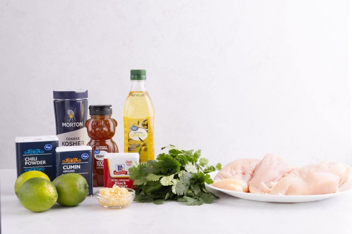 Chili powder, limes, cumin, salt, honey, black pepper, garlic, oil, cilantro, and chicken on a white countertop.