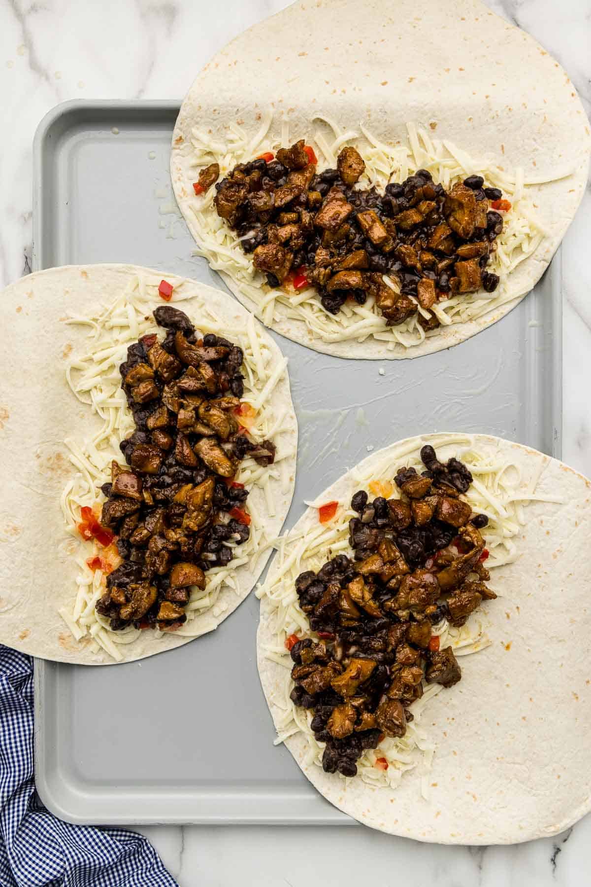 Tortillas laid flat on a sheet pan with quesadilla filling spread on half of each one.
