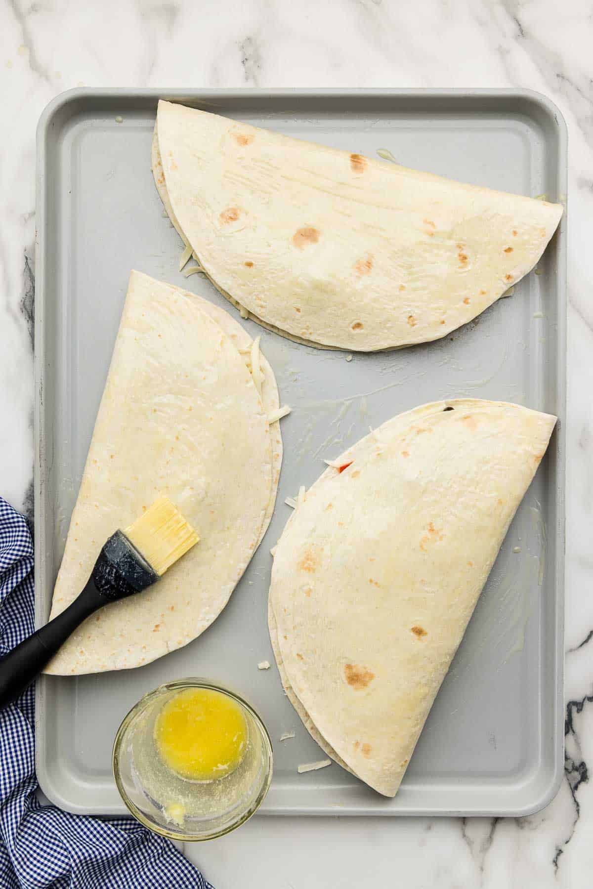 Folded quesadillas on a pan with a silicone brush and a small dish of butter.