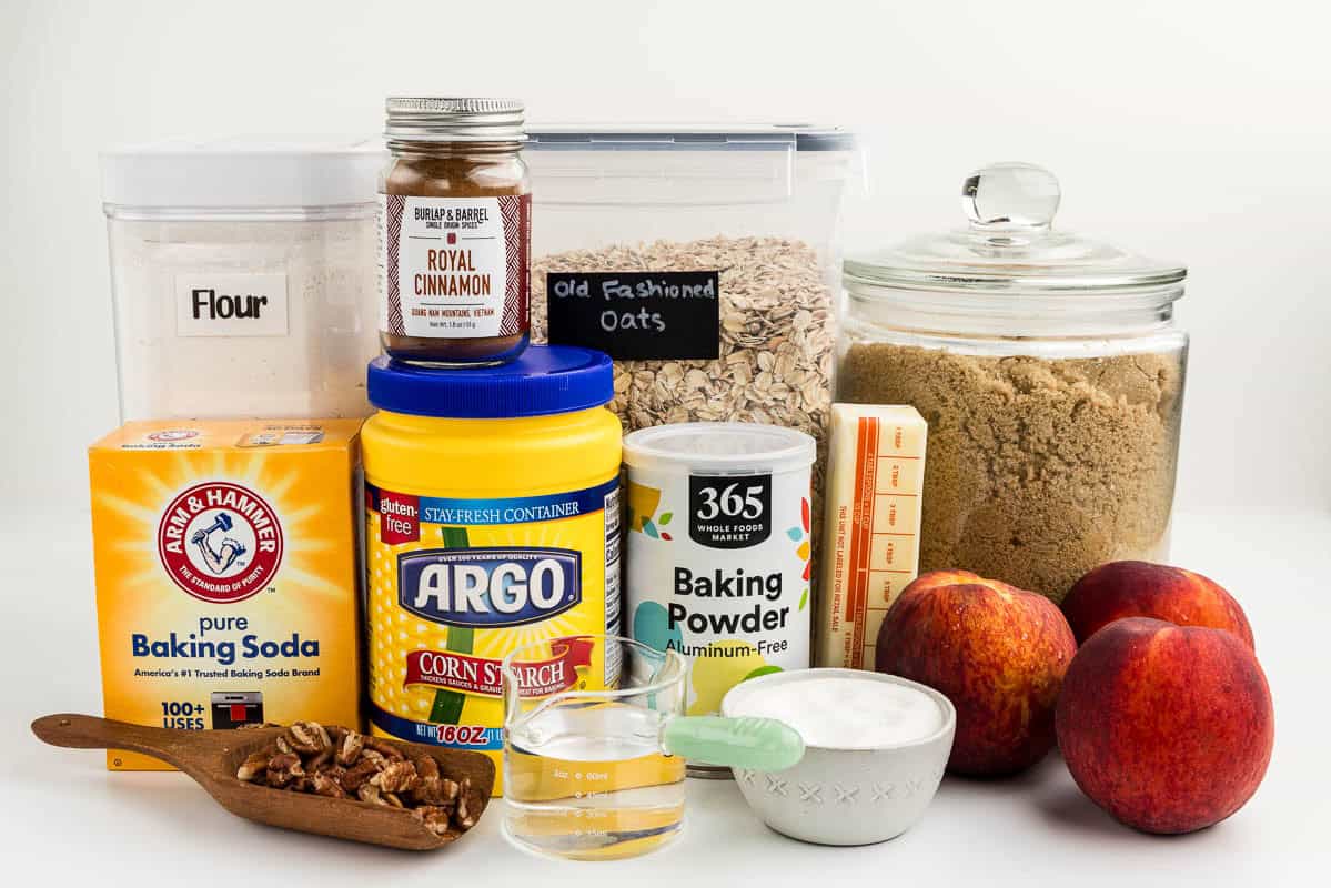Flour, nuts, water, cinnamon, corn starch, baking powder, oats, butter, peaches, brown sugar, and baking soda on a white surface.