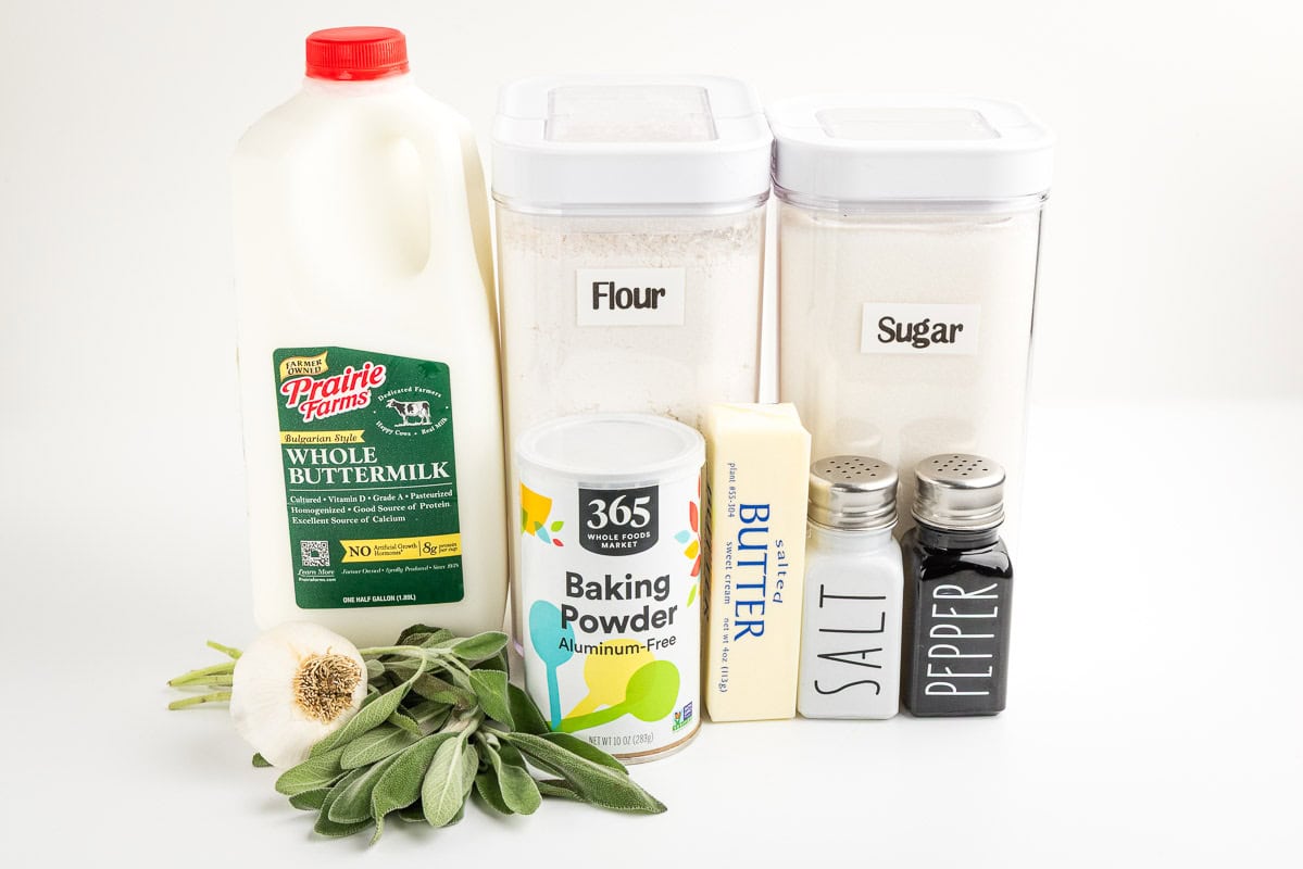 Garlic, sage, buttermilk, baking powder, flour, sugar, butter, salt, and pepper on a white surface.