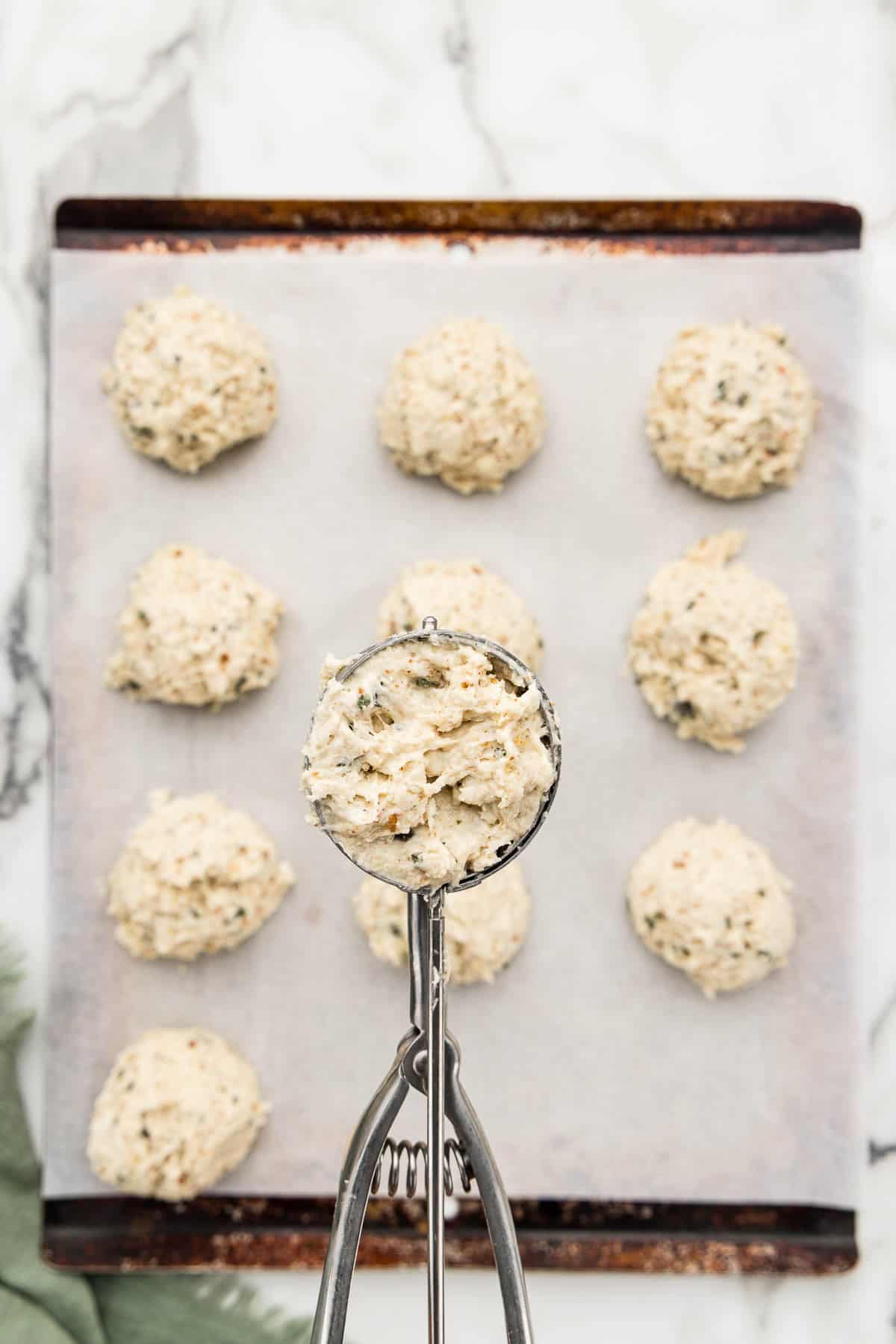 A ball of dough in a cookie scoop over a pan with rows of raw biscuits.