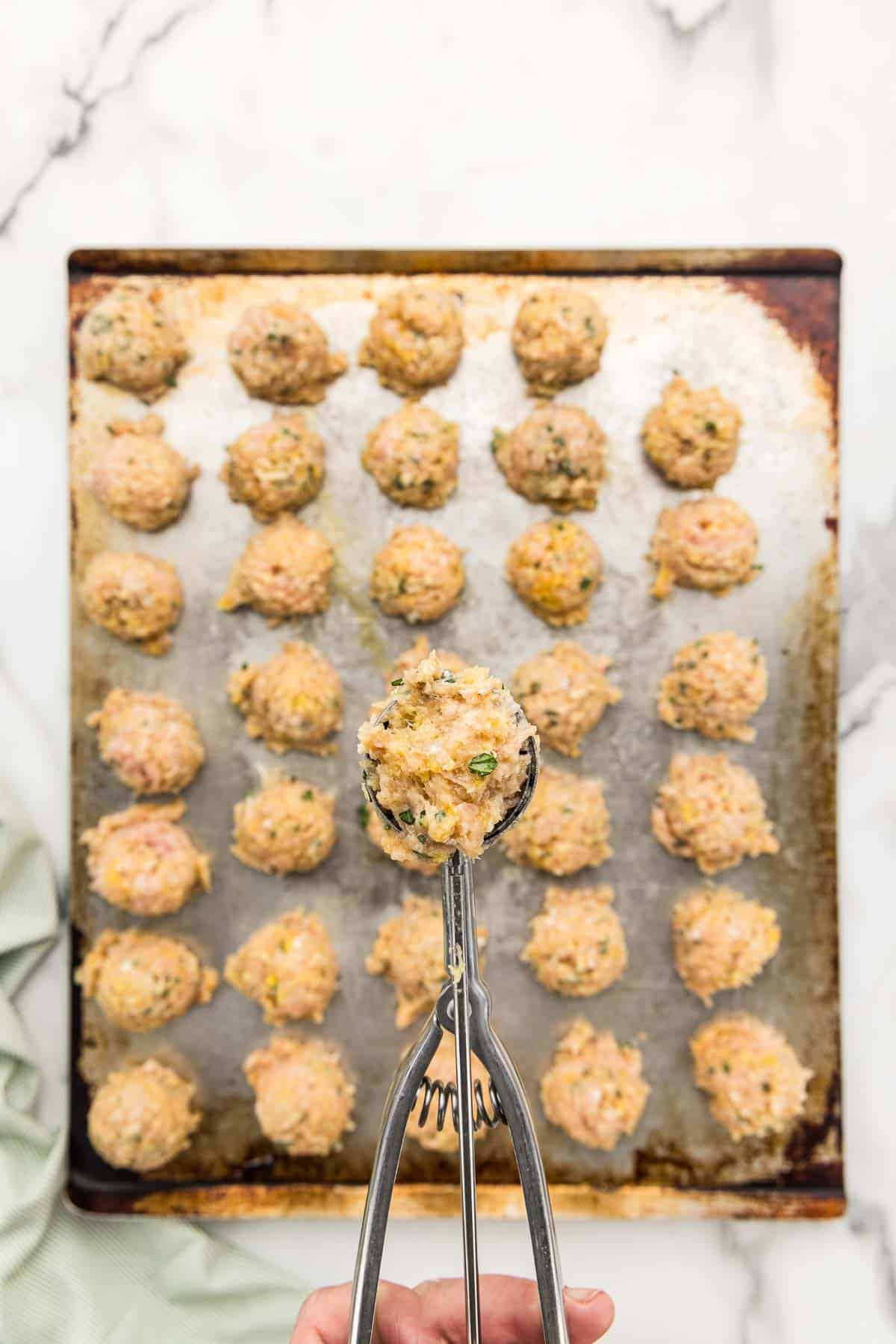 A cookie scoop holding a raw meatball over a sheet pan lined with meatballs.