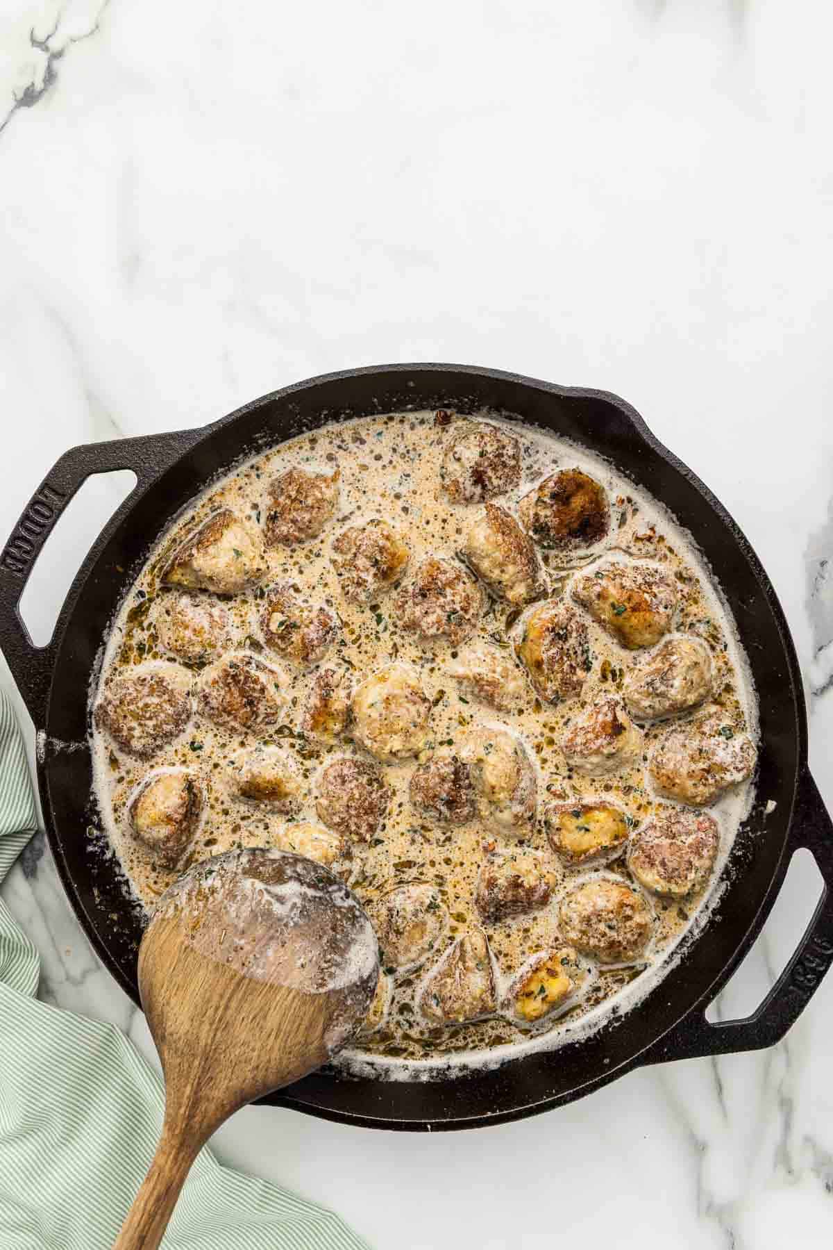Meatballs in sauce cooking in a skillet with a wooden spoon.