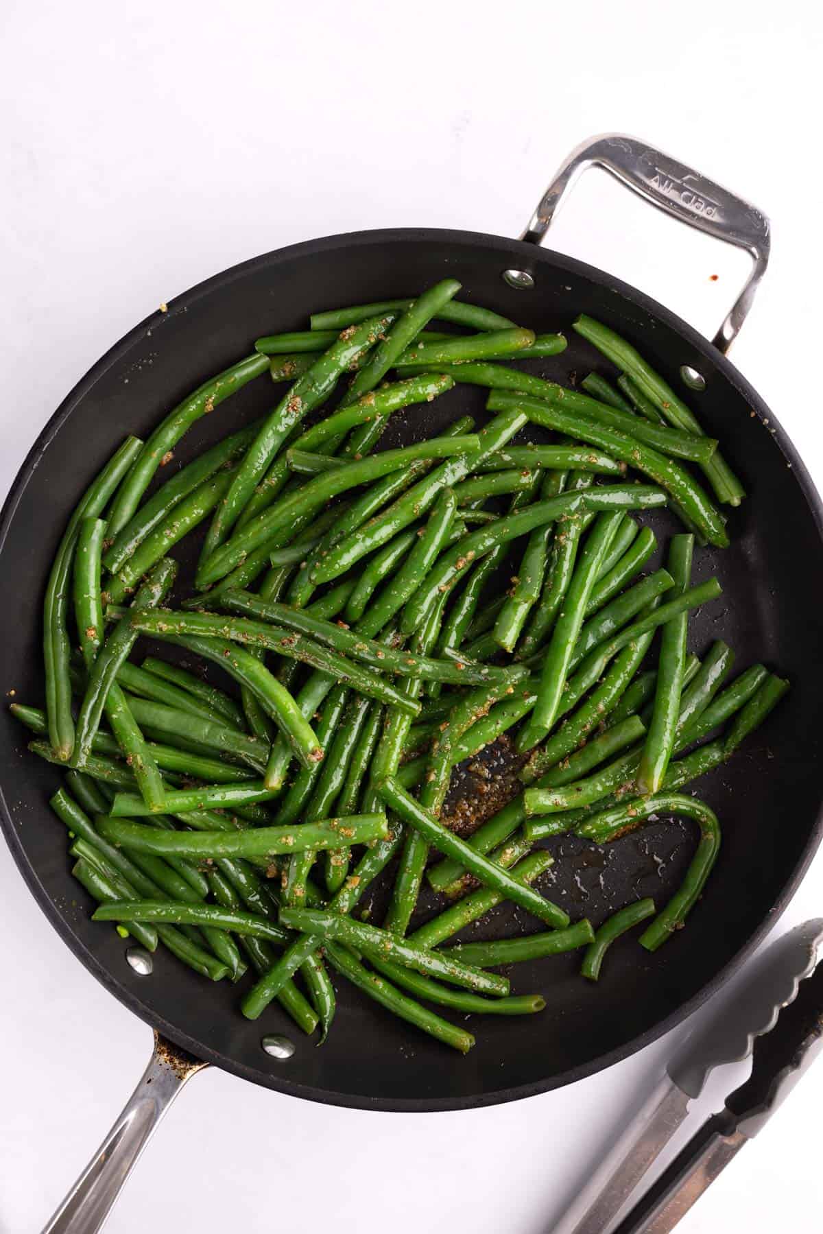 Green beans sprinkled with seasoning in a pan with a pair of tongs.